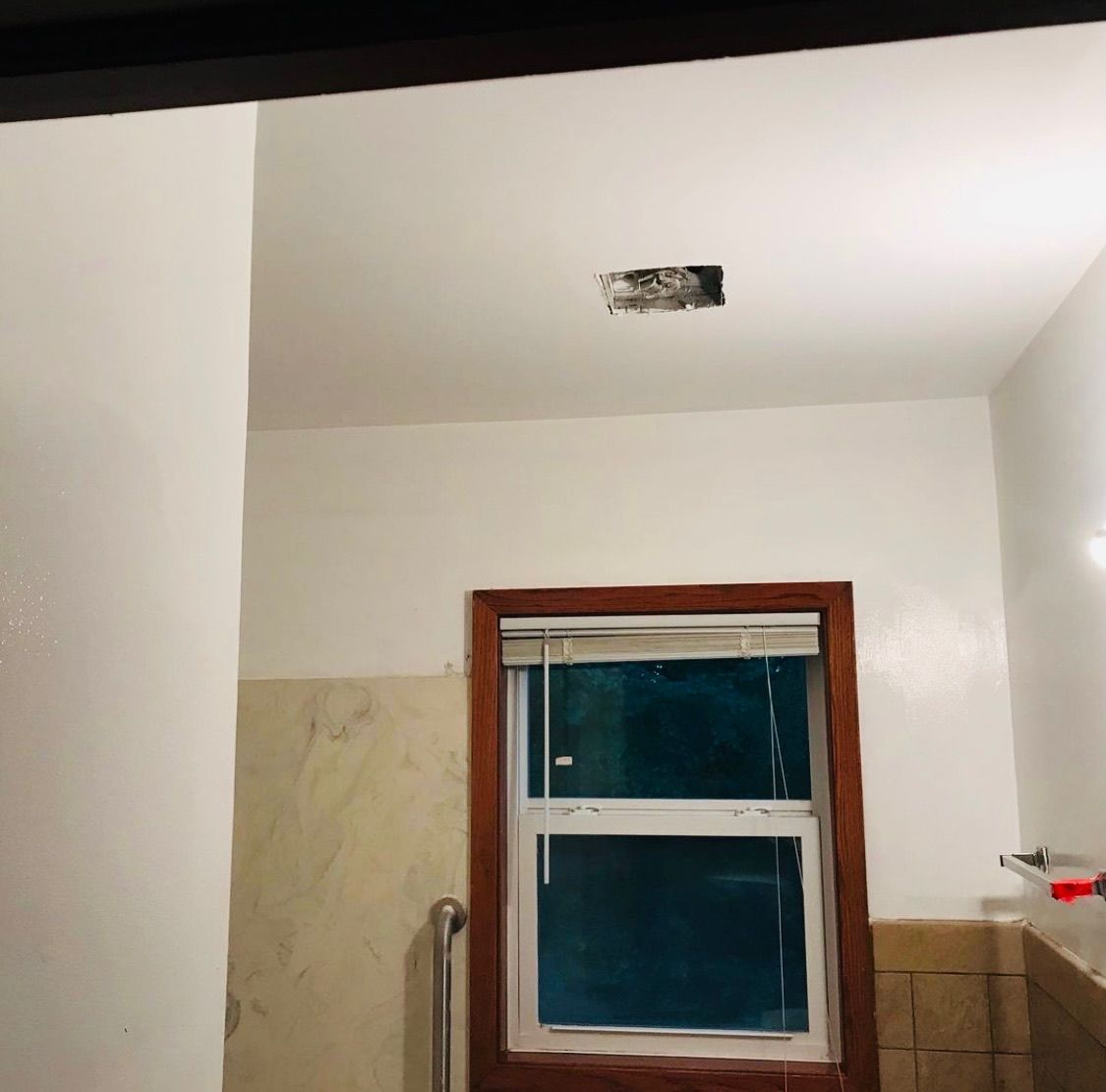 A bathroom interior showing a window with a dark wooden frame, partially tiled walls, and a hole in the white ceiling.