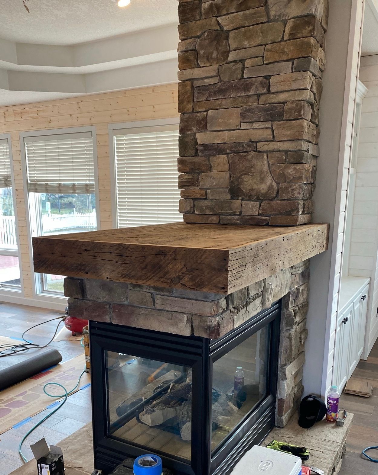 A fireplace in a room under construction, featuring a natural stone facade and a thick, rustic wooden mantle.