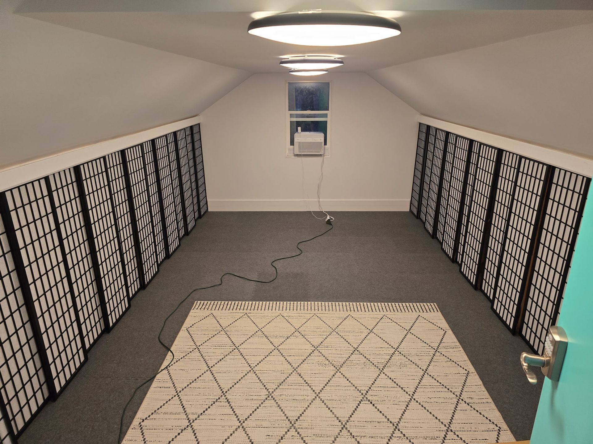 Attic room with sloped ceilings, rows of binary code panels on walls, a rug, ceiling lights, and a window air conditioner.