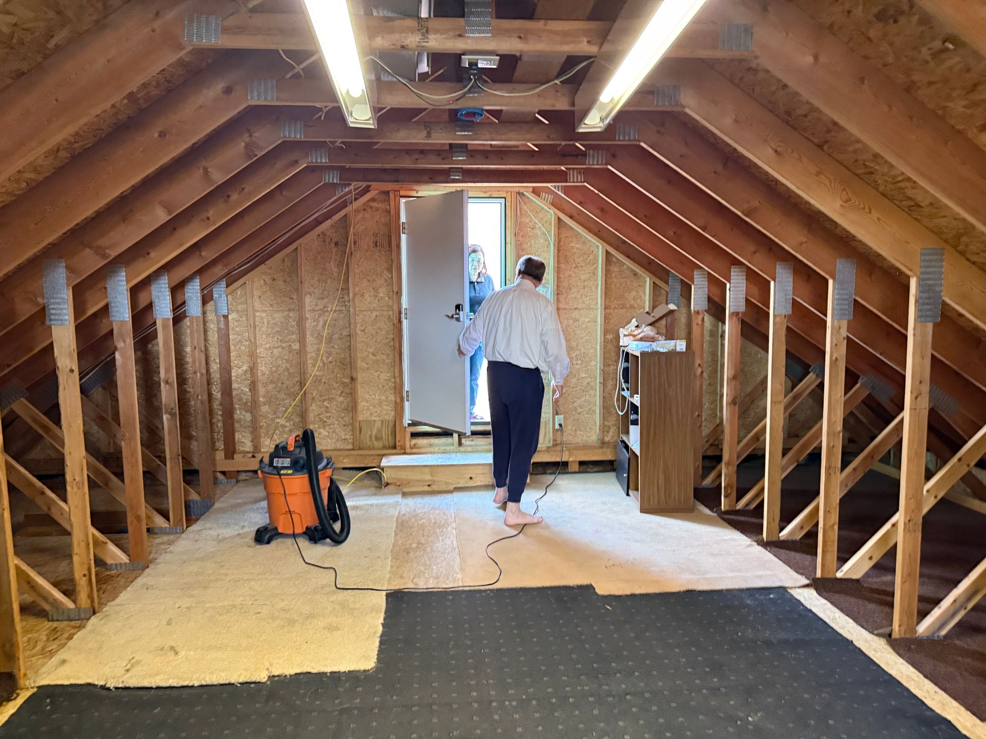 A person stands in a wooden attic with an open door at the end, a shop vacuum on the floor, and unfinished paneling.