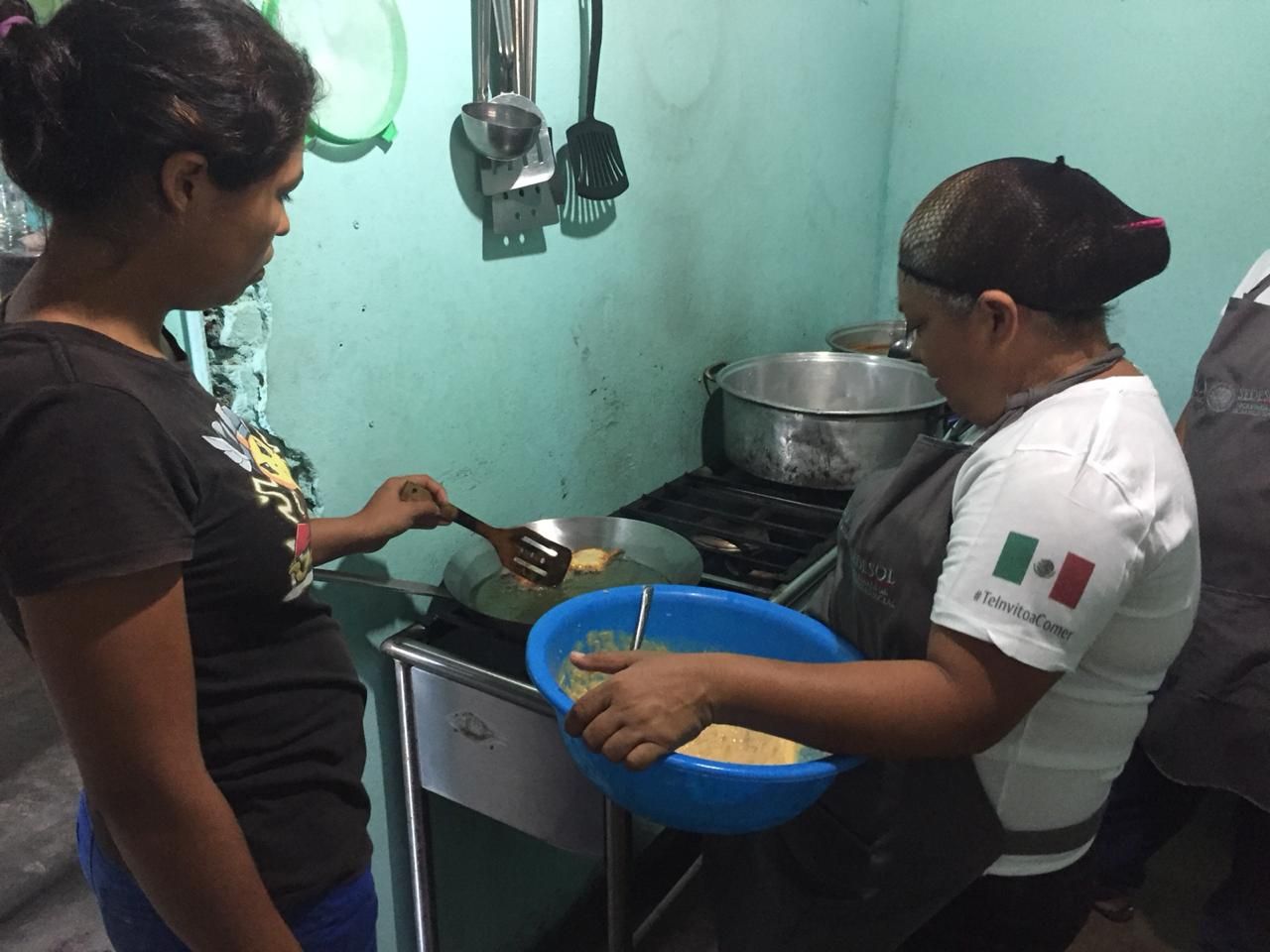 Two people washing dishes in a small kitchen, one holding a blue basin beside a sink and stove.
