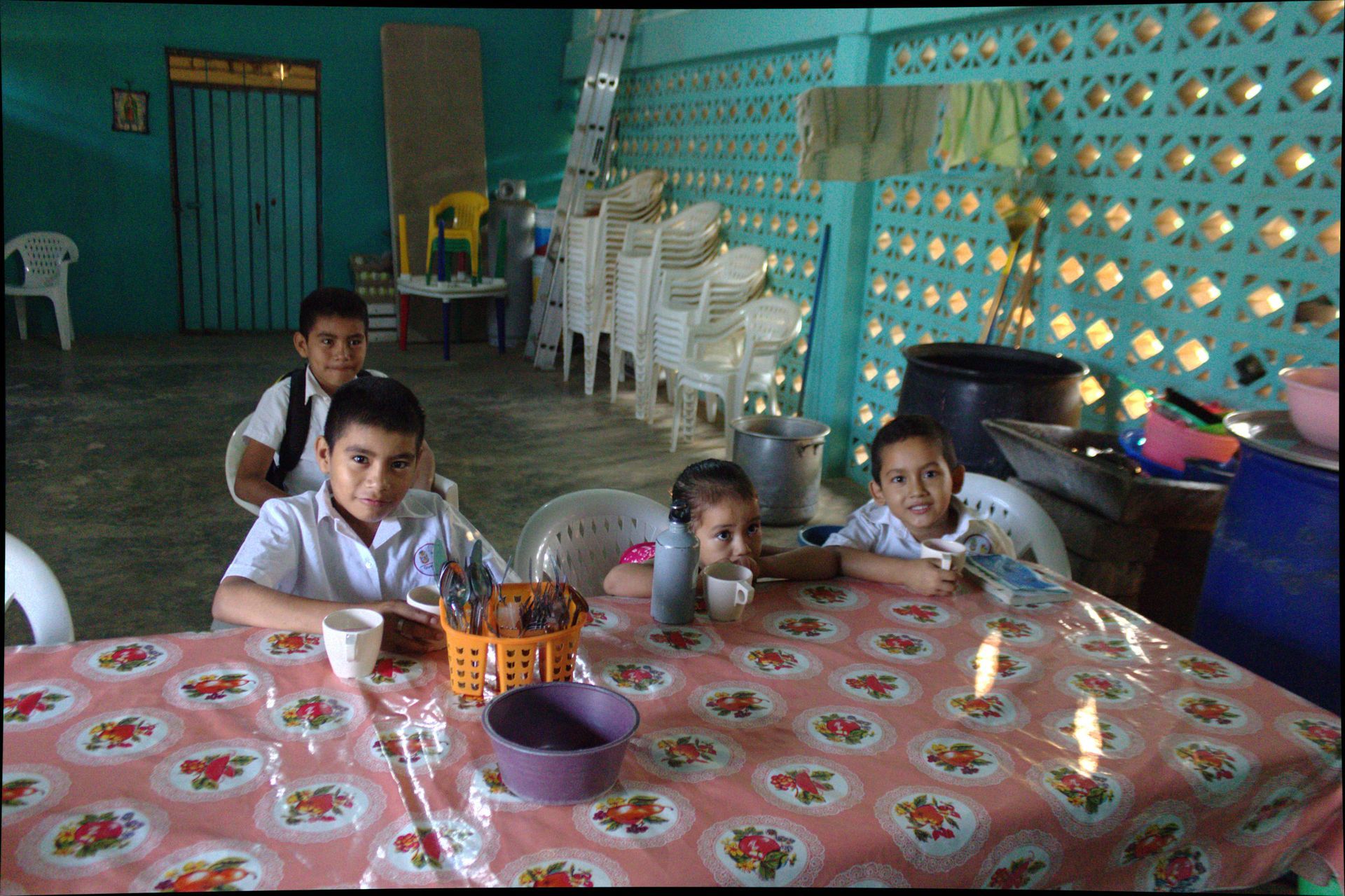 Children sit at a table in a turquoise cafeteria, eating with cups and plates.