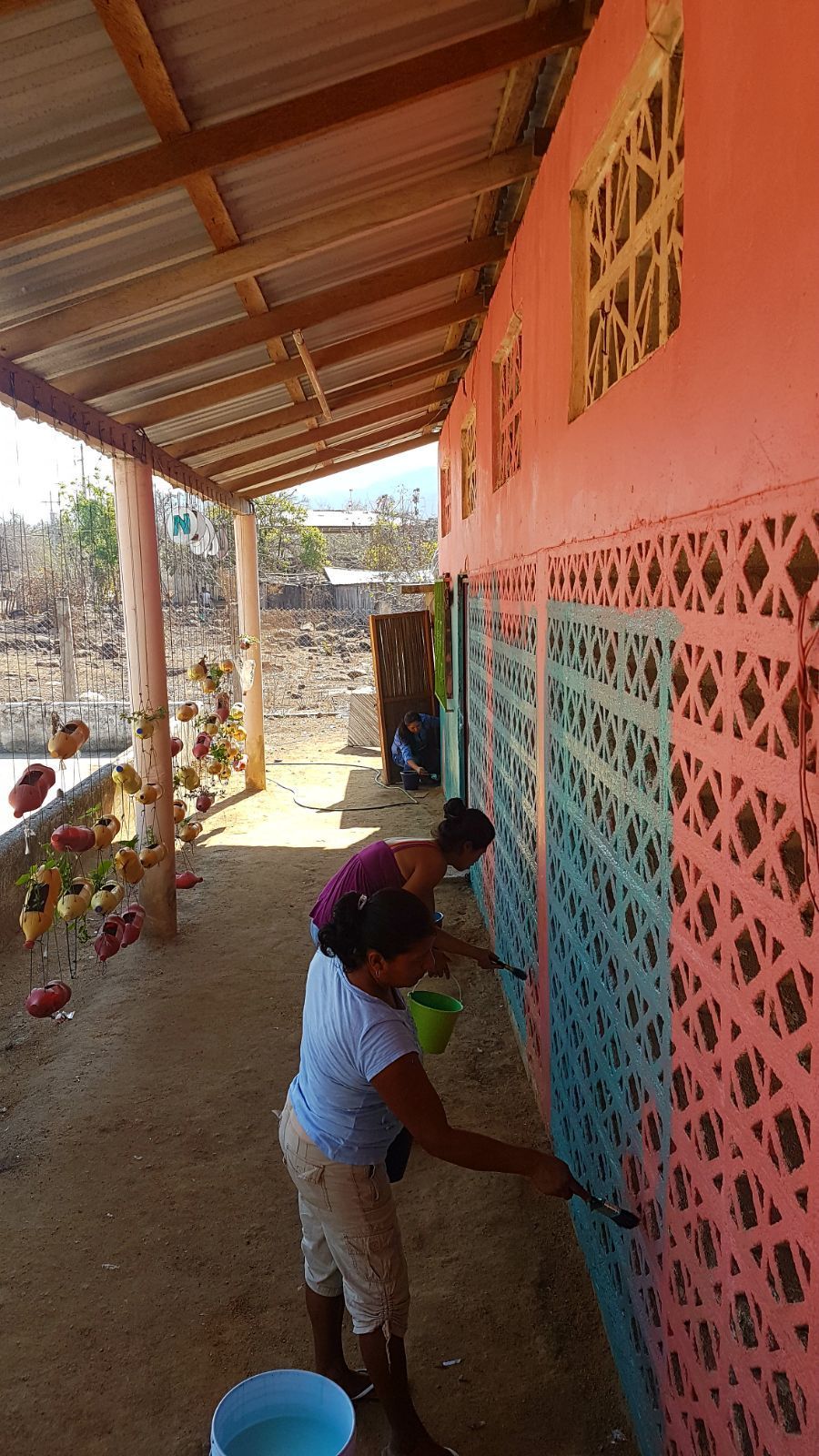 Person painting a pink perforated wall on a covered porch beside a coral-colored building.
