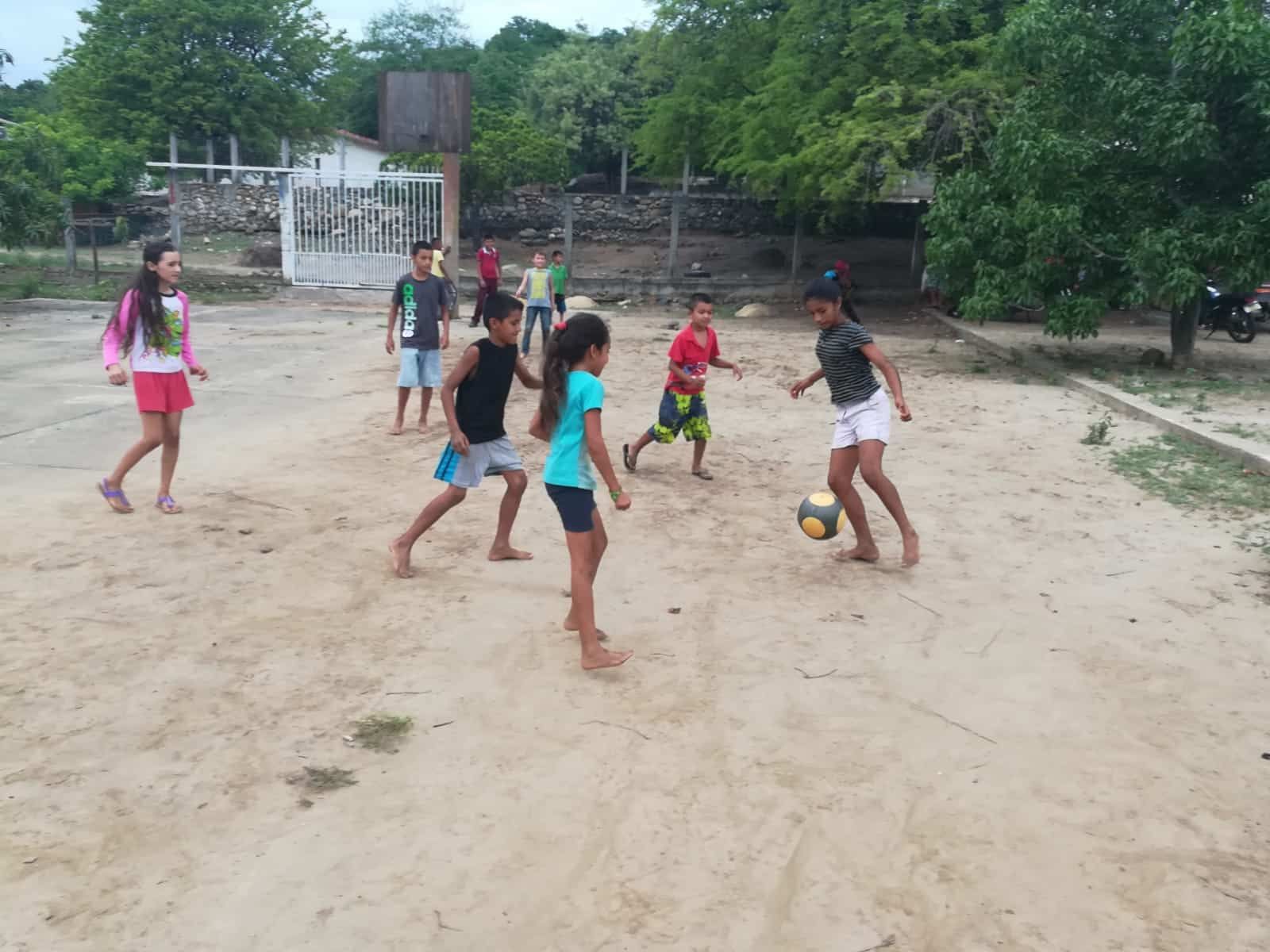 Children playing soccer on a dirt field near a goal and trees