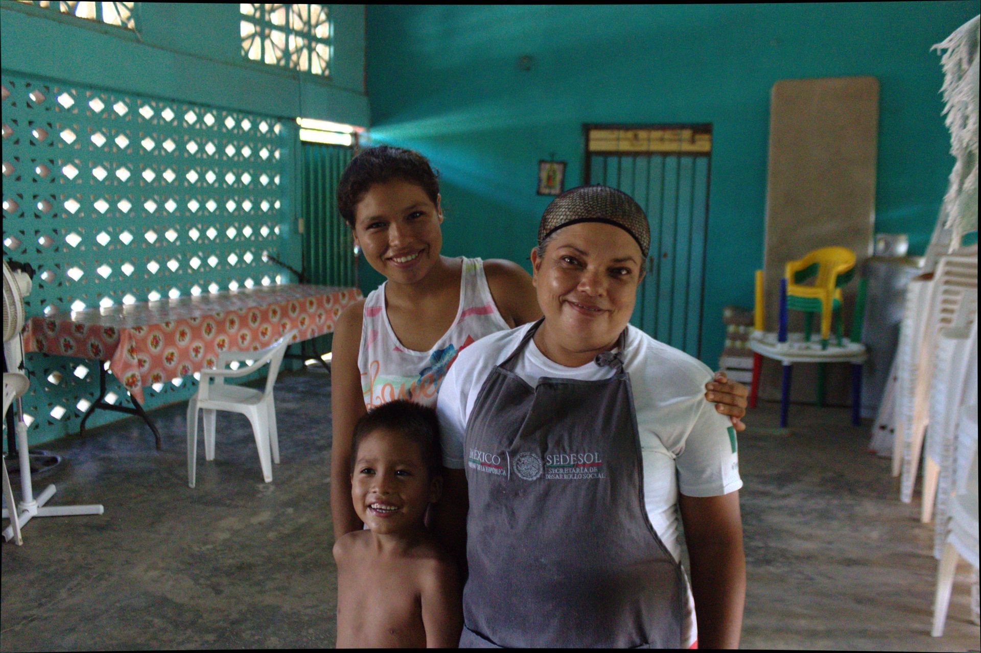 Three people stand in a teal-painted room with plastic chairs and tables in the background.