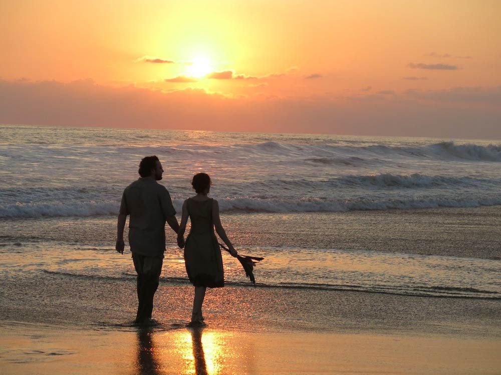 Two people walking hand in hand on a beach at sunset, with waves and orange sky.