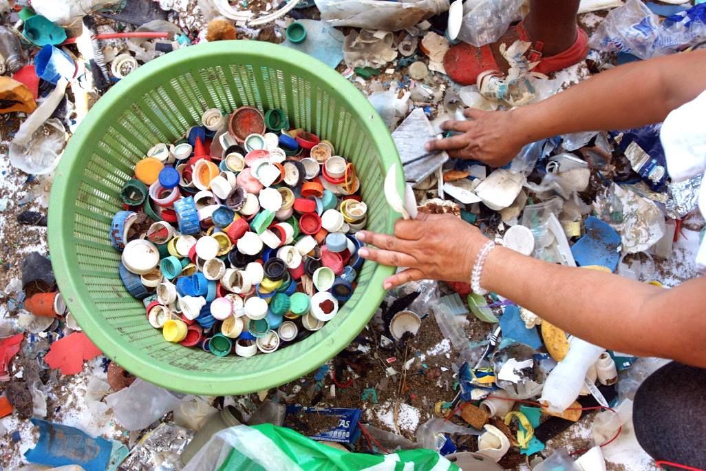 Hands sorting colorful plastic bottle caps into a green basket amid mixed trash.