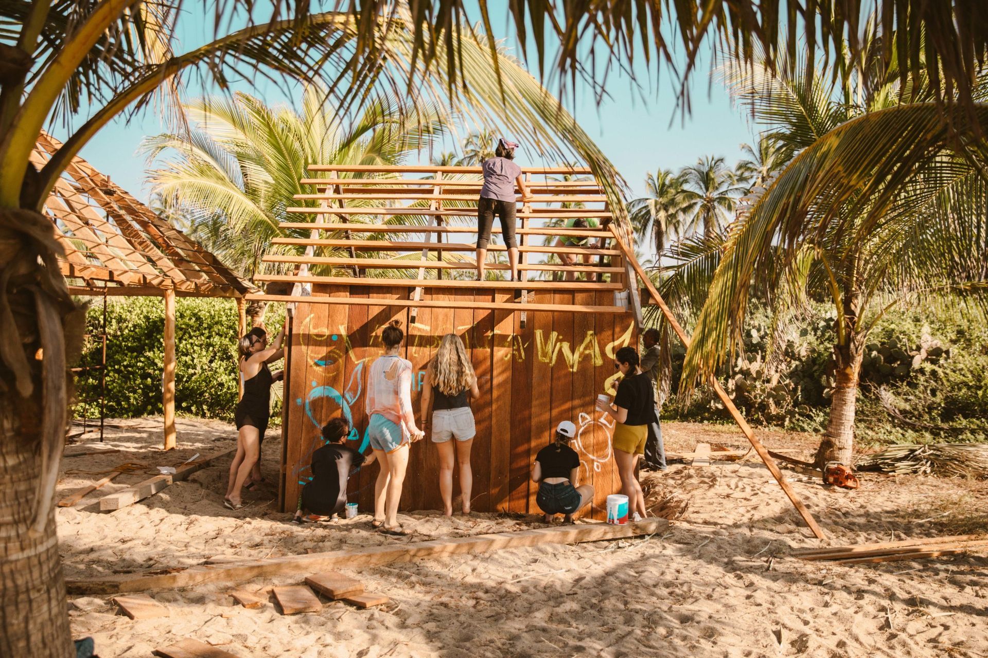 People on a beach under a wooden hut frame, with palm trees and a person on the rooftop.