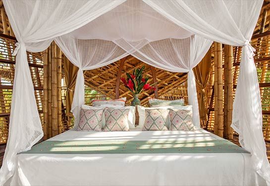 Canopy bed with white drapes, patterned pillows, and tropical flowers in a rustic bamboo hut