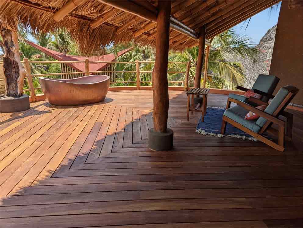 Wooden deck with a hammock, chairs, and a stone tub under a thatched roof, overlooking tropical palms