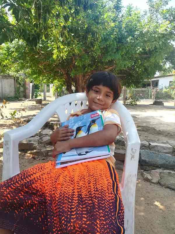 Child smiling in a white chair outdoors, holding books and a cup, with trees and a yard in the background