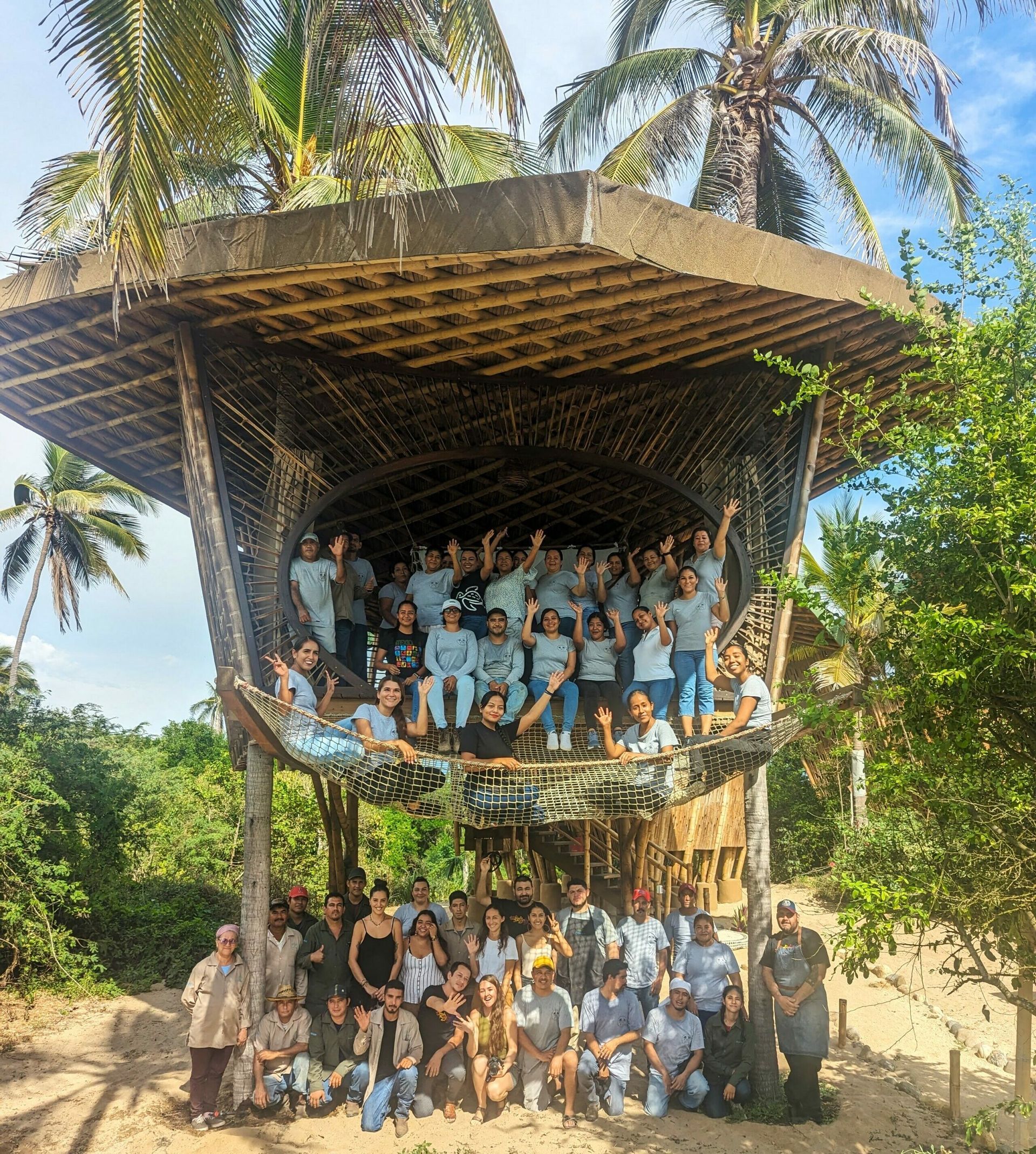 Large group posing in a two-level open wooden structure under palm trees in bright daylight