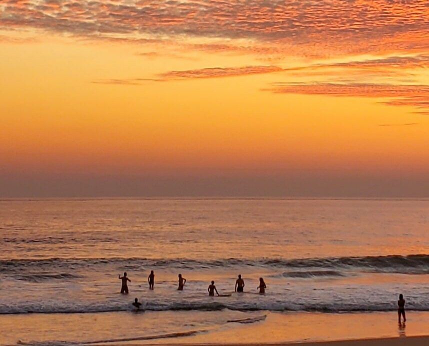 Sunset over calm ocean with small silhouettes of people wading near the shore