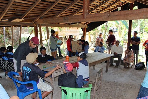 People gathered under a thatched pavilion for a community meeting, seated around tables in a rural setting.