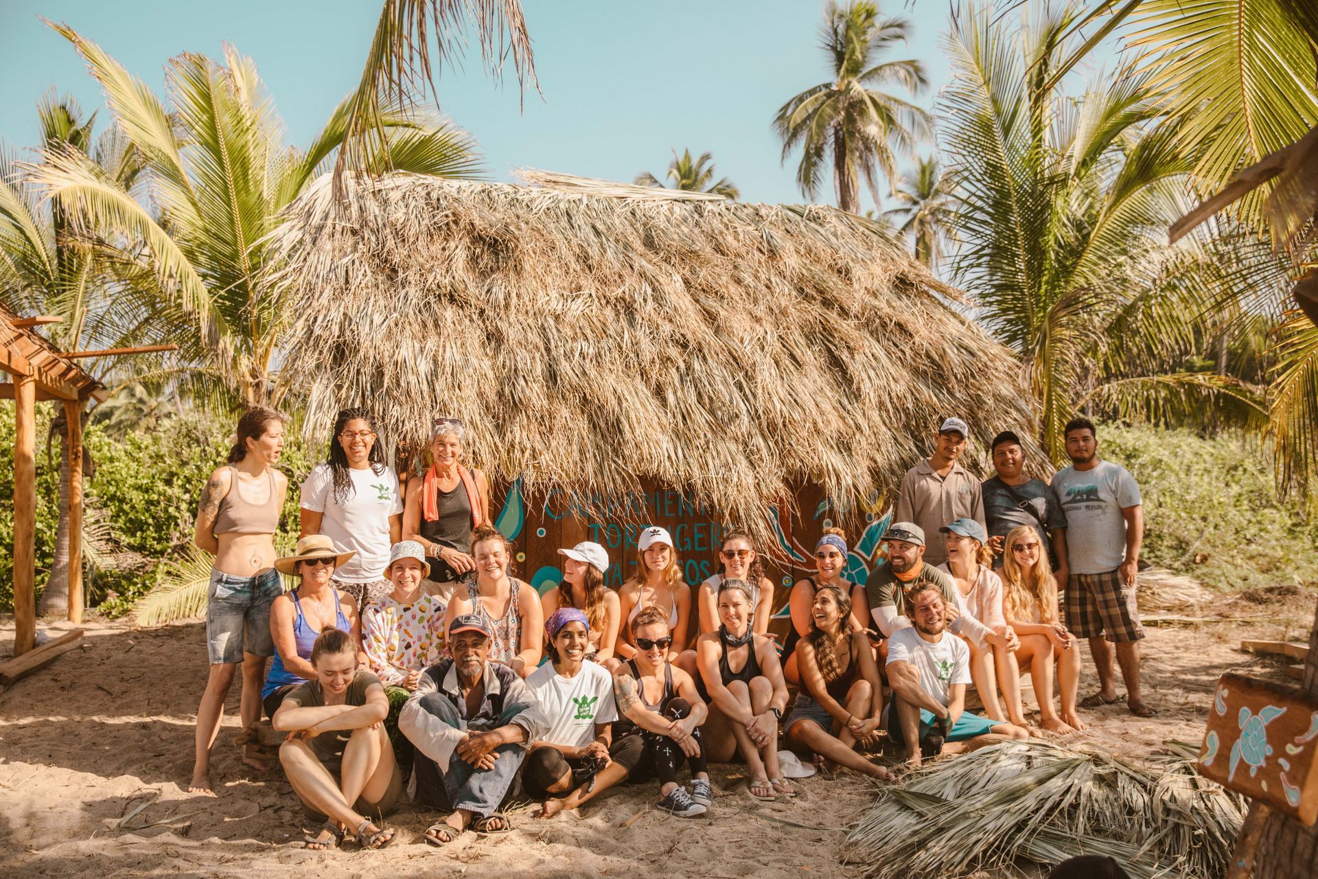 Group posing outside a thatched hut on a sunny tropical beach with palm trees