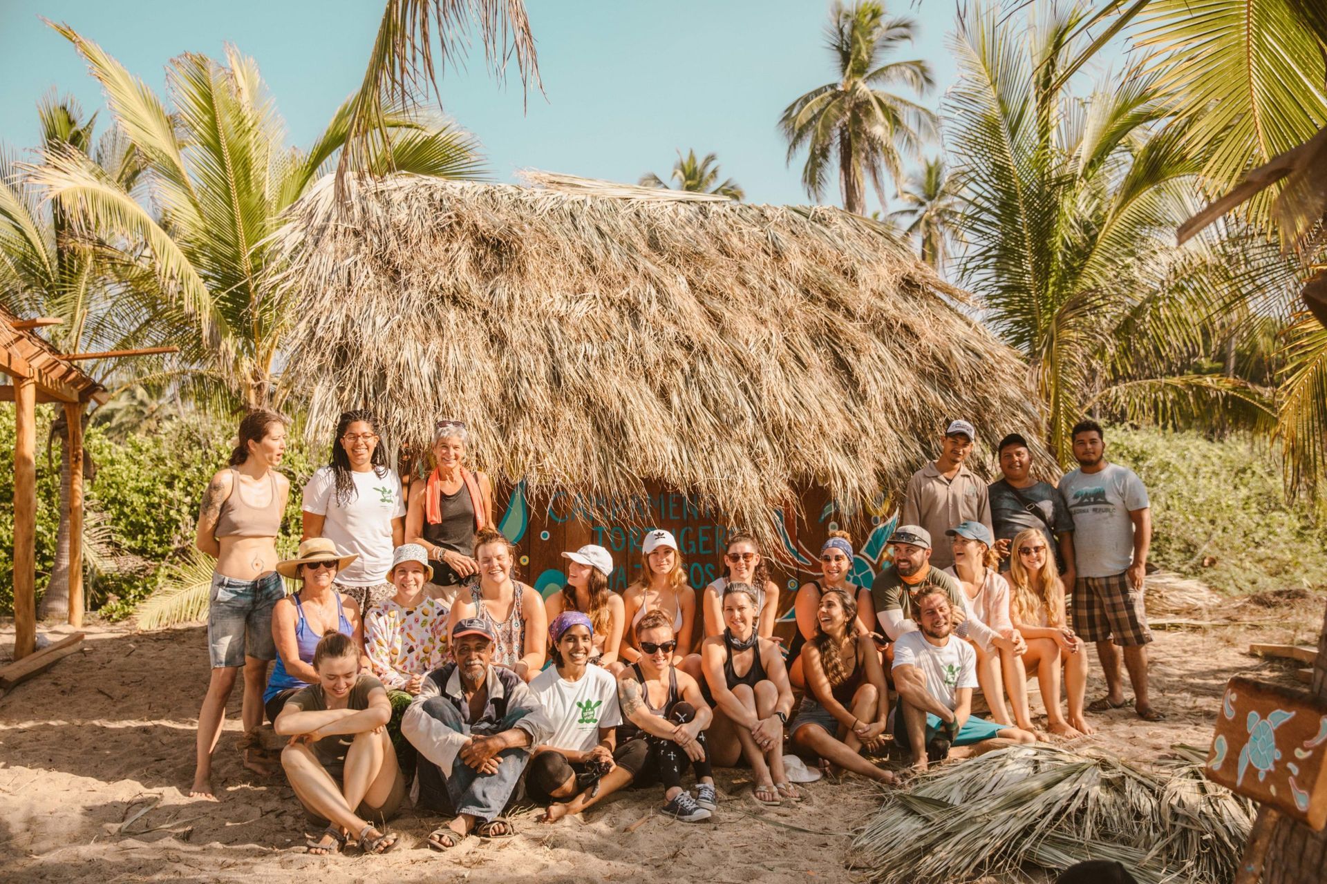 Group posing in front of a thatched hut with palm trees on a sunny tropical beach