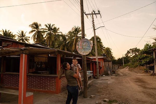 Two people walking beside a dirt road past red buildings and palm trees at sunset