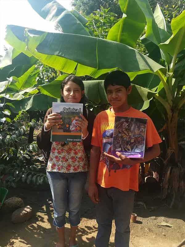 Two people standing outdoors in front of large banana leaves, each holding a colorful book or box.