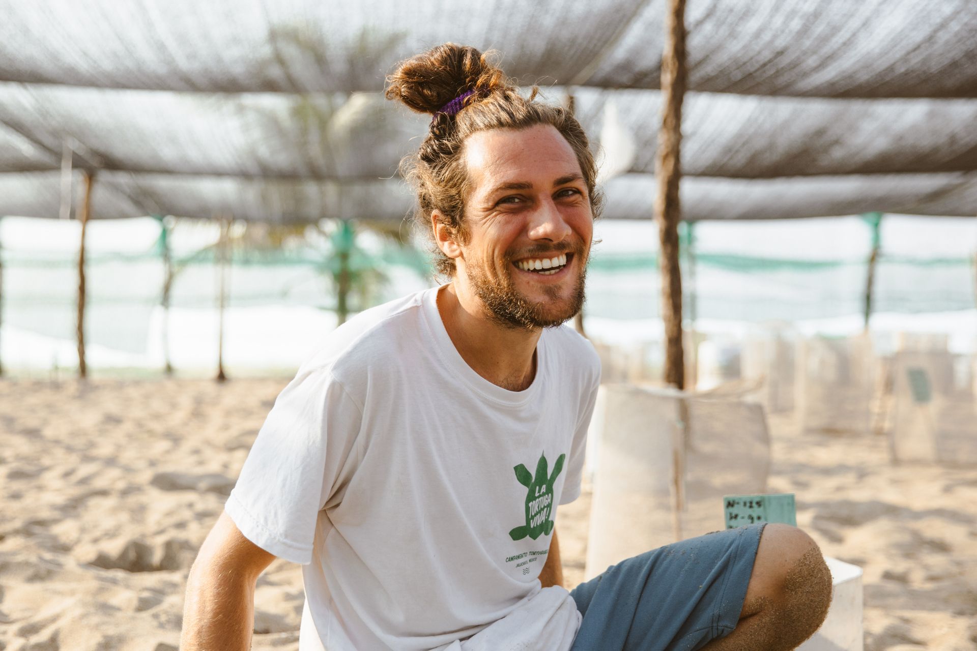 Smiling person in a white T-shirt sitting in a greenhouse with rows of potted plants
