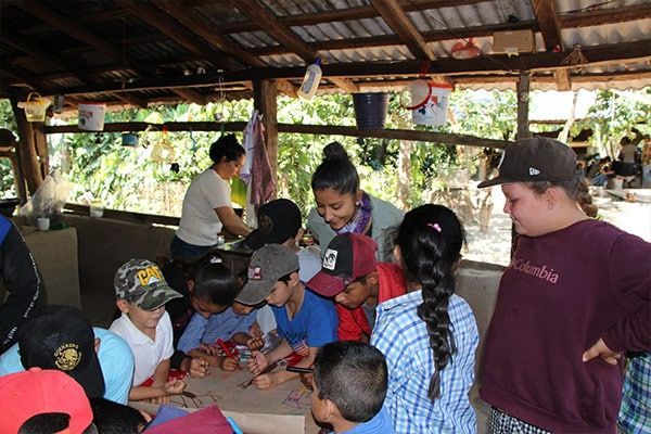 Children gathered around a table in a covered outdoor pavilion, watching an activity with a woman nearby.