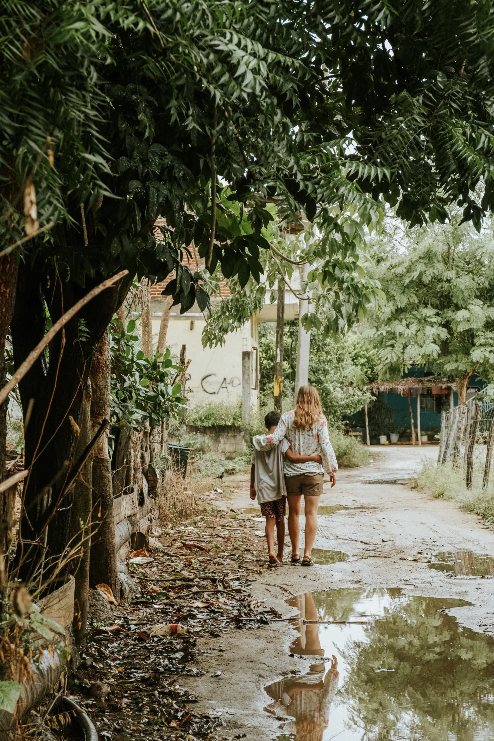 Two people walking along a muddy path beside trees, with puddles reflecting them.