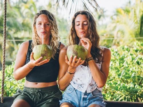 Two people drinking from coconuts on a tropical porch, with greenery in the background