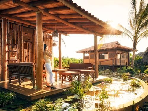 Sunlit wooden porch by a tropical pond, with a person standing beside bamboo furniture and palm trees.