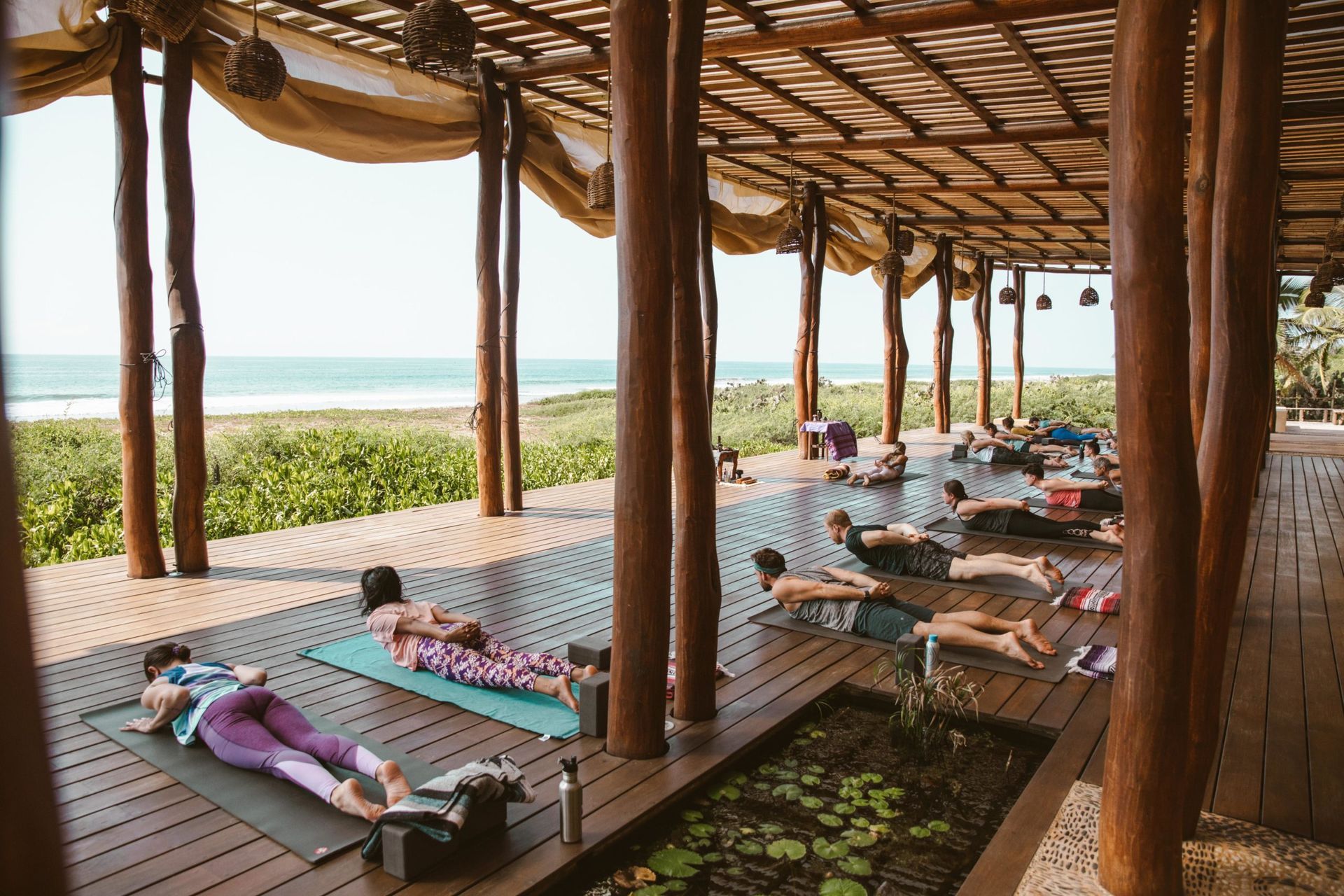 People lying on yoga mats under a seaside pavilion, with ocean view beyond.