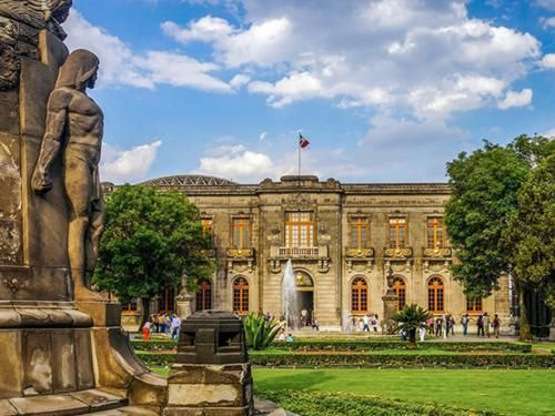 Historic palace facade with a fountain, statue, and people in a landscaped garden under a blue sky