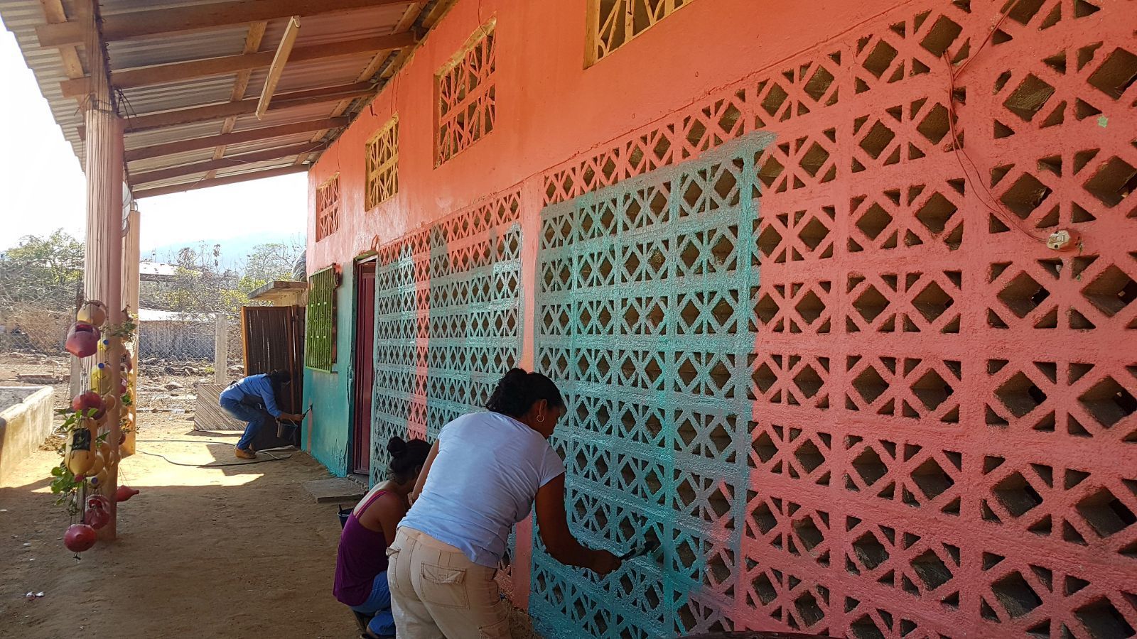Two people repainting a pink and turquoise school wall with decorative concrete blocks under a roofed walkway