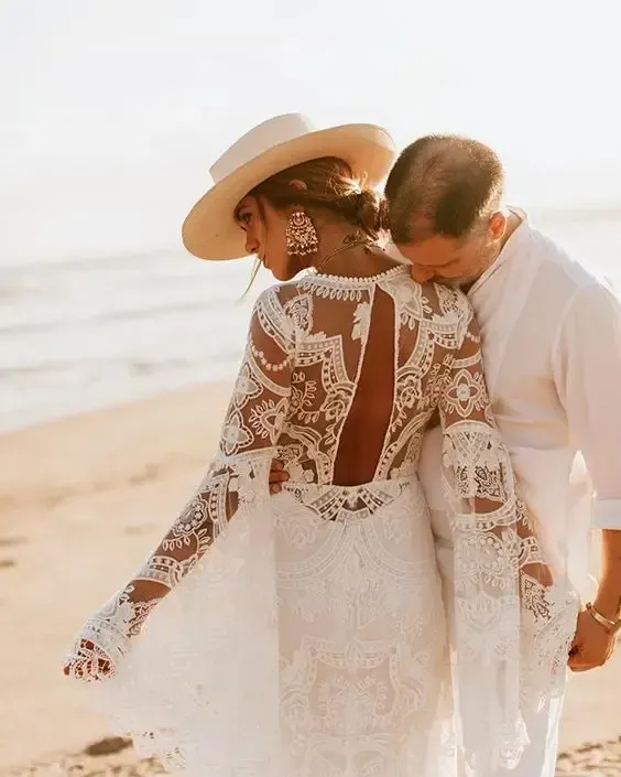 Couple in white wedding attire embracing on a sunny beach, with lace dress and wide-brim hat