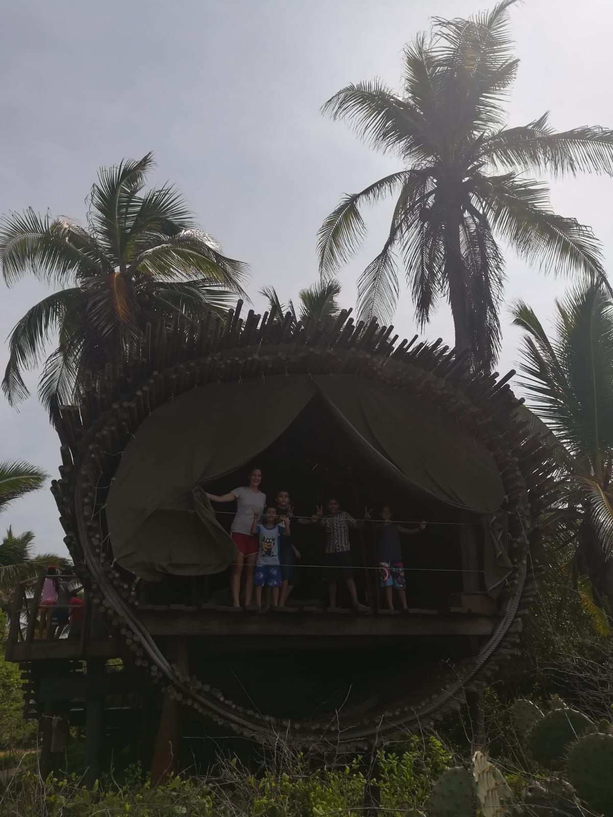 People standing inside a large circular palm-frond hut with palm trees around it.