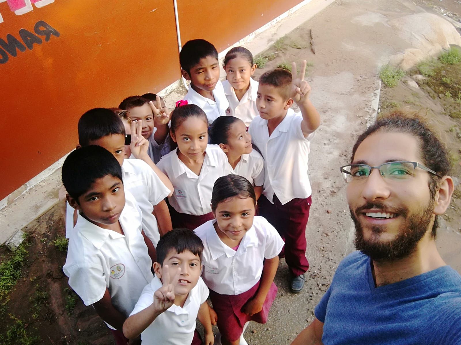 Group of students in white uniforms posing with a smiling teacher in front of an orange wall