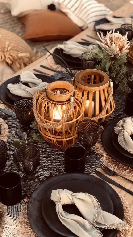Cozy table setting with woven candle lanterns, dark dishes, folded napkins, and dried flowers on a neutral cloth.