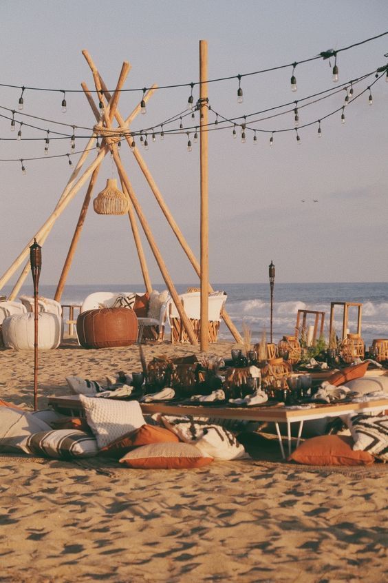 Beach scene with teepee poles, string lights, and cushions beside the sea at sunset