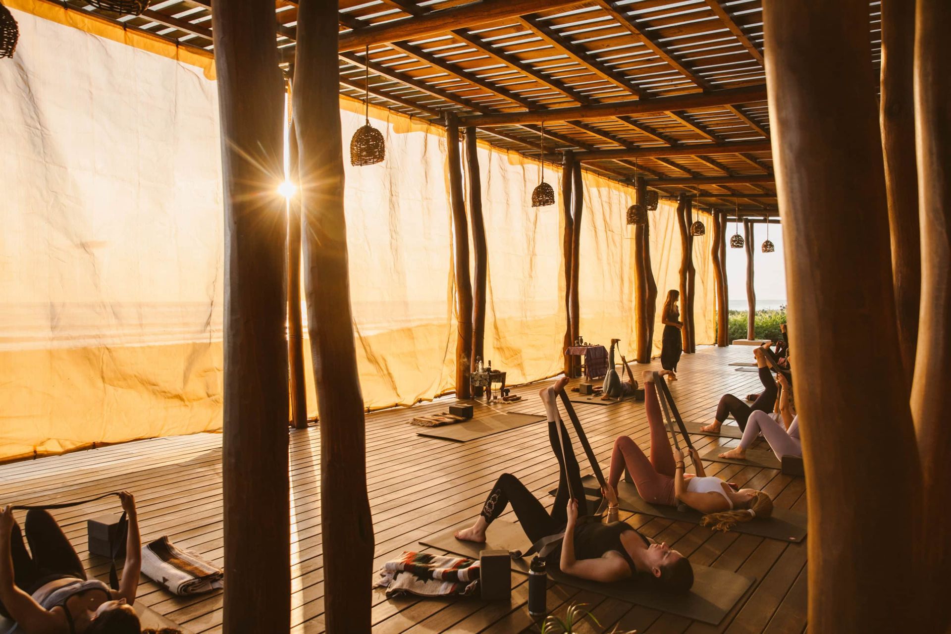 Sunlit rustic shelter with woven mats, wooden posts, and a few lounge chairs on a sandy floor