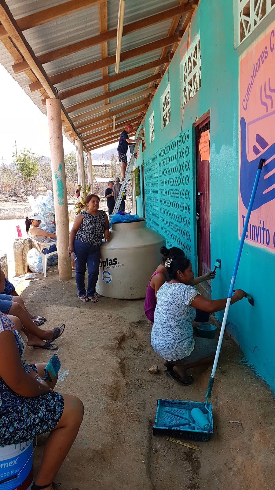 People painting a turquoise wall under a covered walkway, with buckets and cleaning tools nearby.