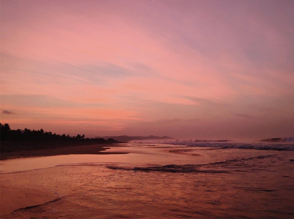 Pink sunset over a calm beach with gentle waves and a dark shoreline silhouette