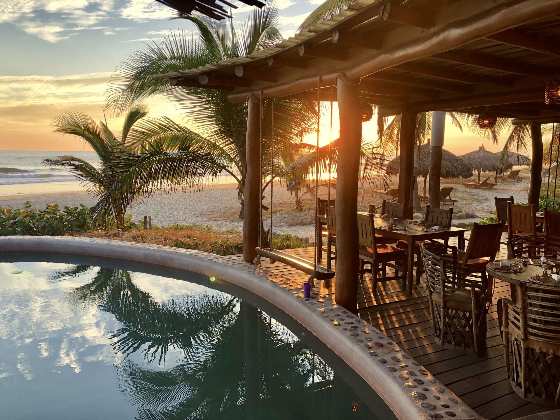 Sunset view from a poolside patio overlooking a beach and ocean, with palm trees and wooden chairs