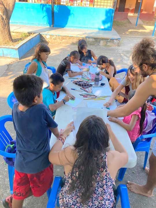 Children gathered around a white table outdoors, doing crafts with colorful materials under blue shade.