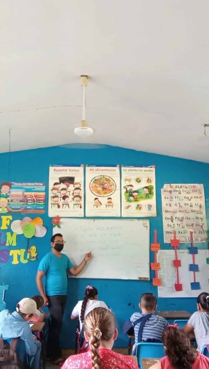 Classroom lesson with teacher pointing at whiteboard, children seated at desks, colorful posters on blue wall