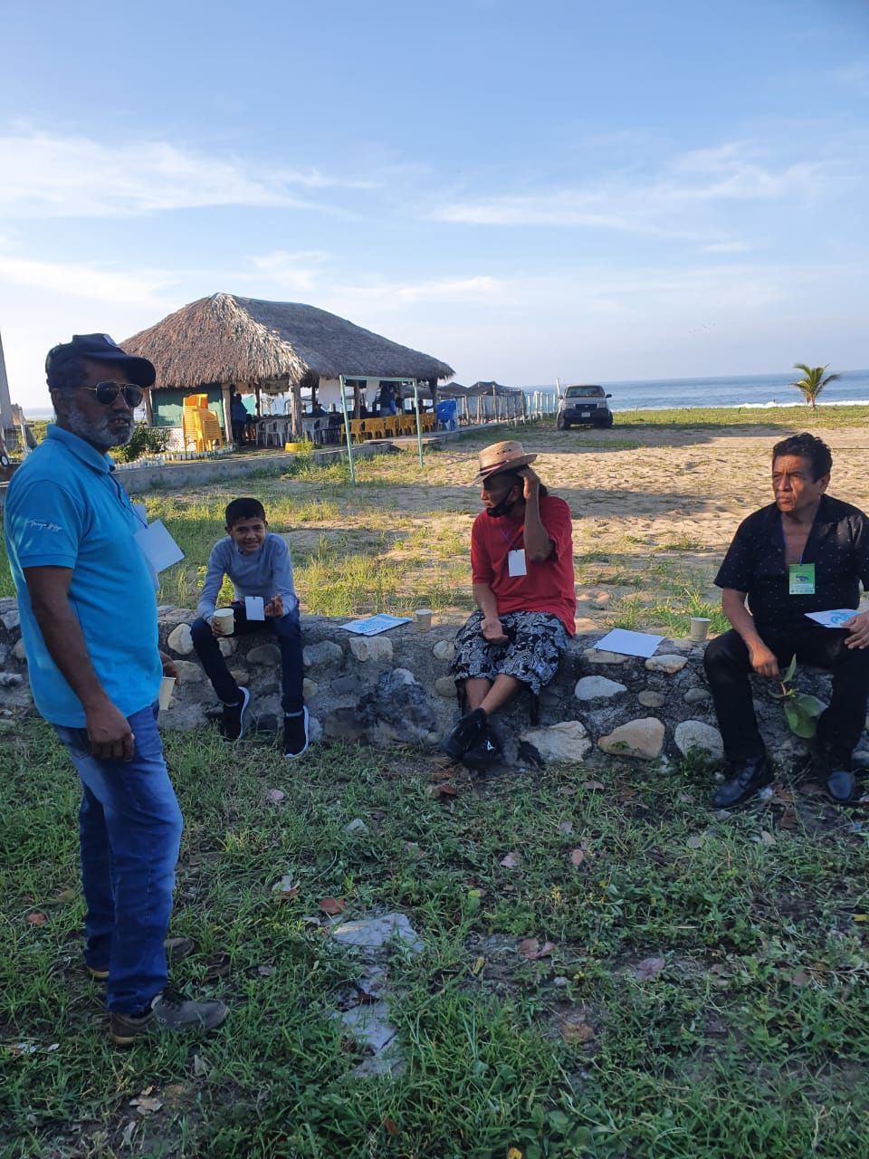People sitting and standing outdoors near a thatched hut by the water at sunset.