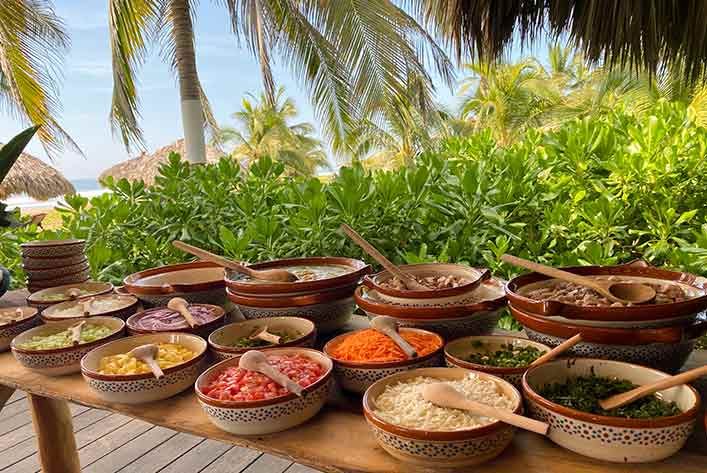 Colorful bowls of prepared food on a wooden table outdoors by tropical palm trees
