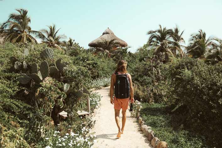 Person walking a sandy path through lush tropical greenery toward thatched huts under palm trees