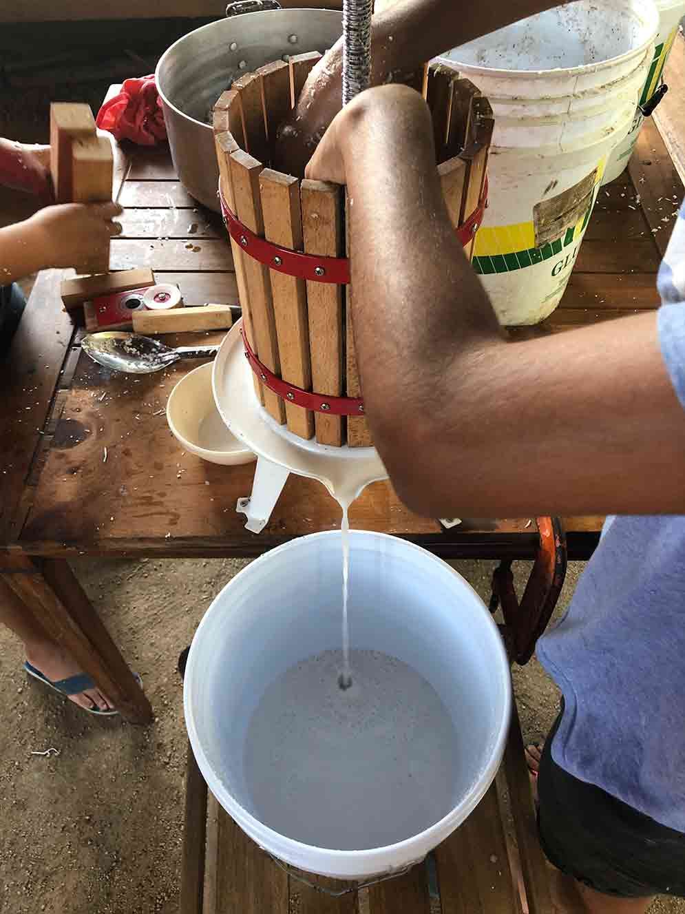 Person pouring liquid from a wooden bucket into a white plastic bucket outdoors