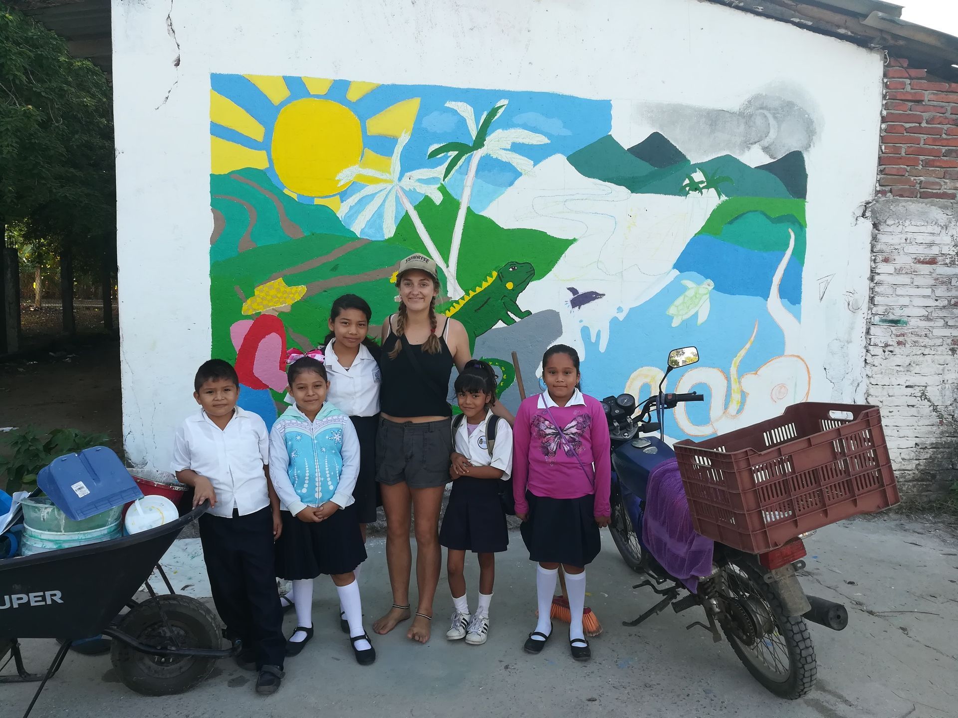 Five children posing before a colorful mural, with bicycles and a cart on either side.