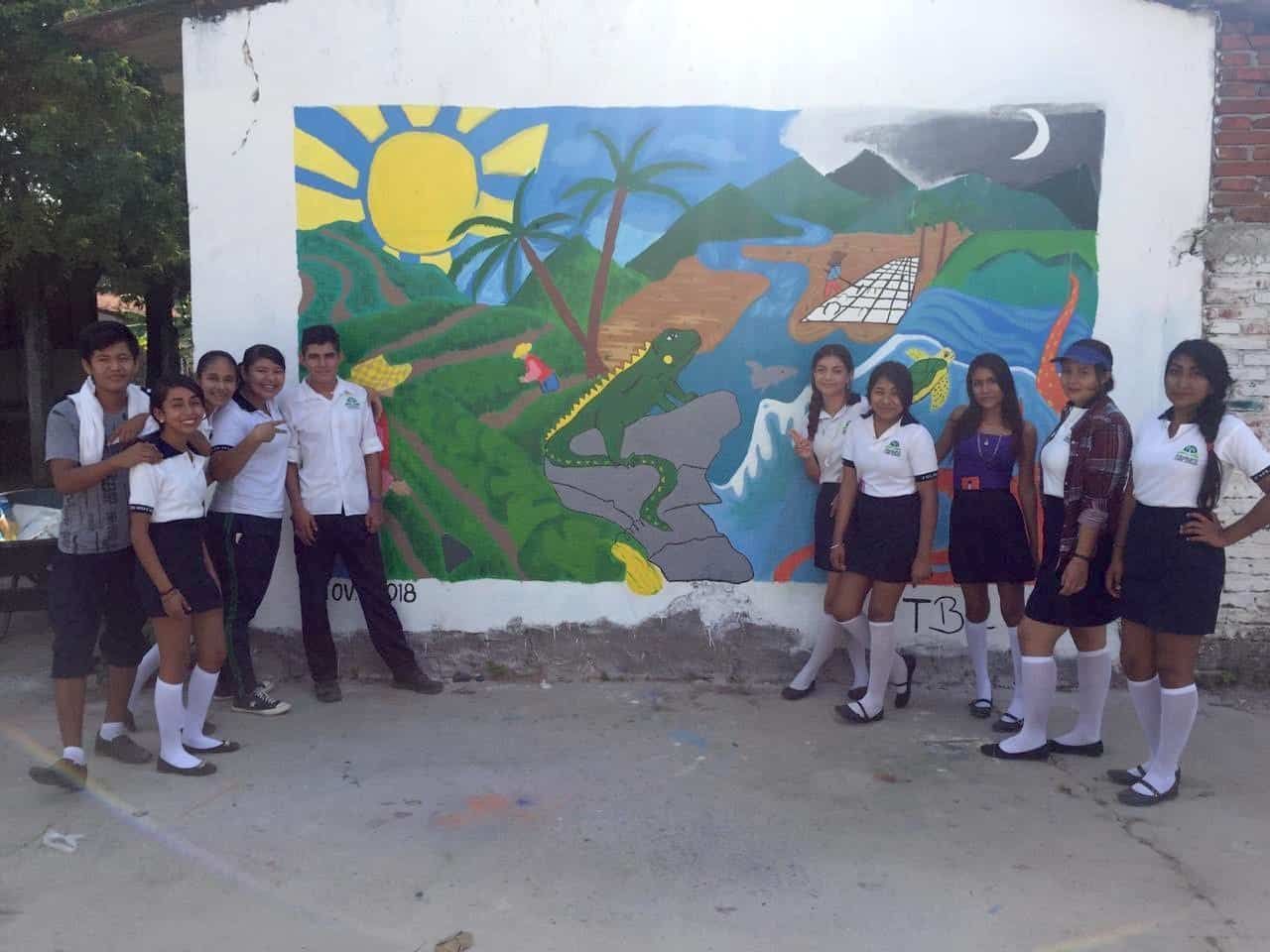 Students pose beside a colorful mural of mountains, sun, moon, and a frog on a wall outdoors.