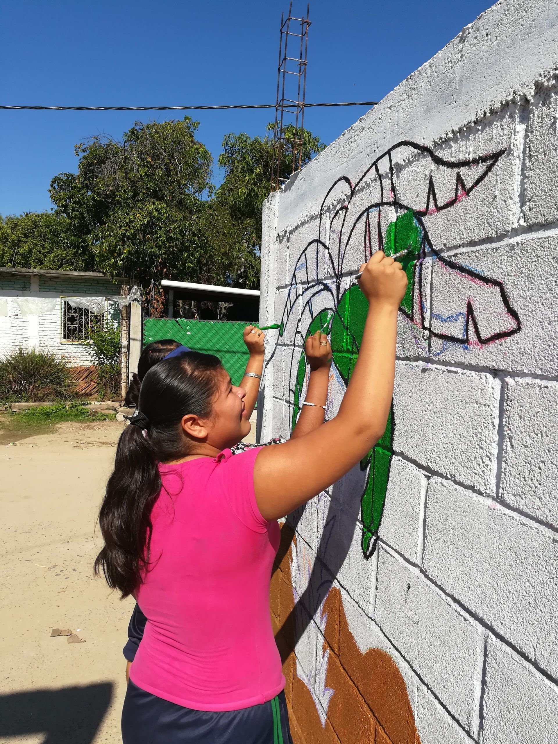 Person painting a mural on a cinder block wall outdoors on a sunny day.