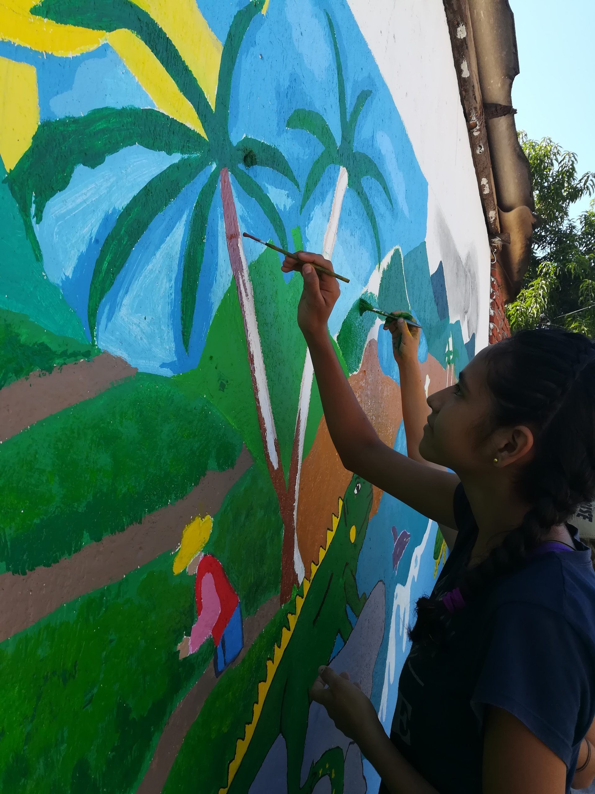 Person painting a colorful mural with palm trees and green hills on an outdoor wall