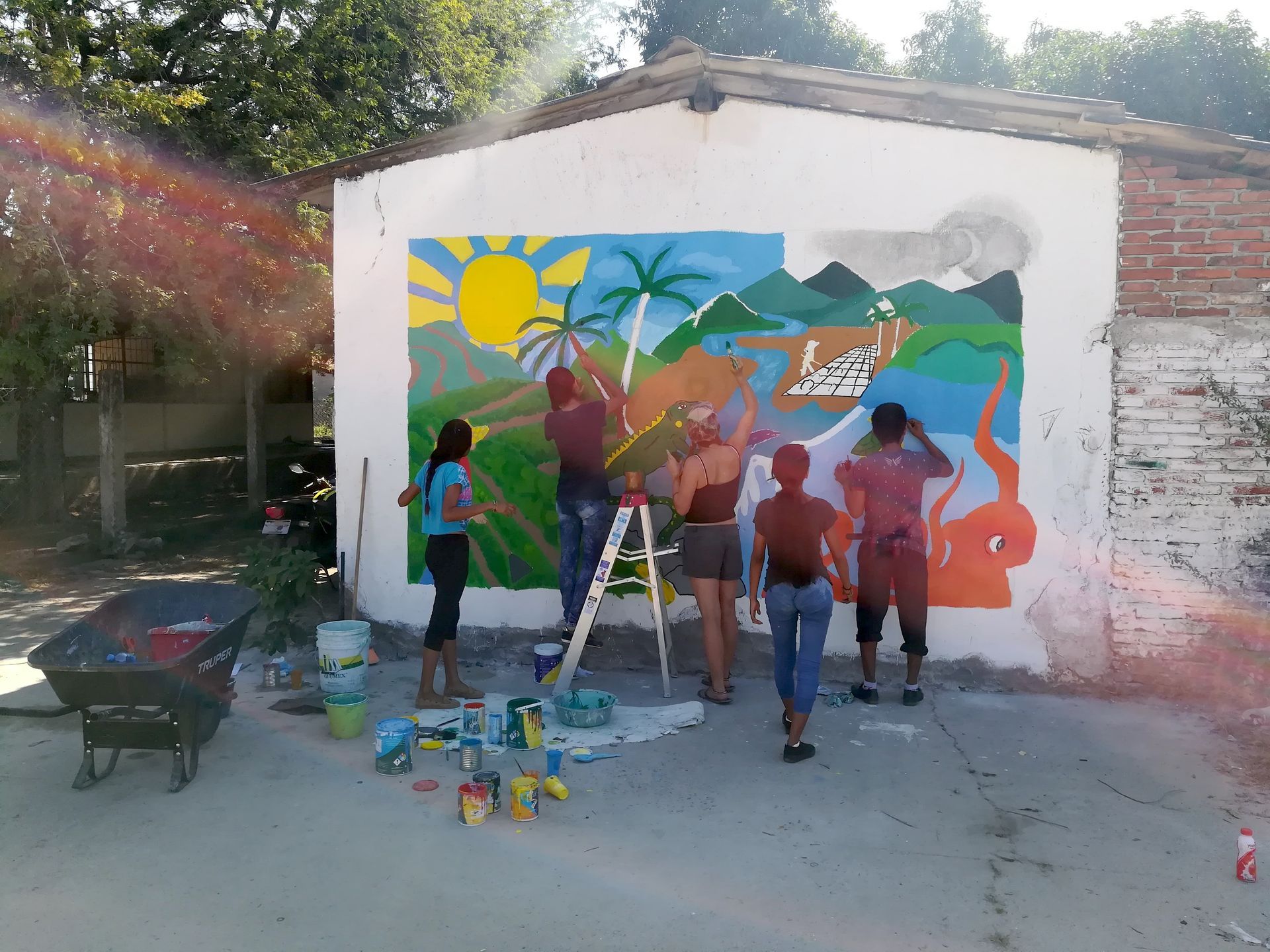 People painting a colorful mural on a white wall outdoors with ladders and paint supplies.